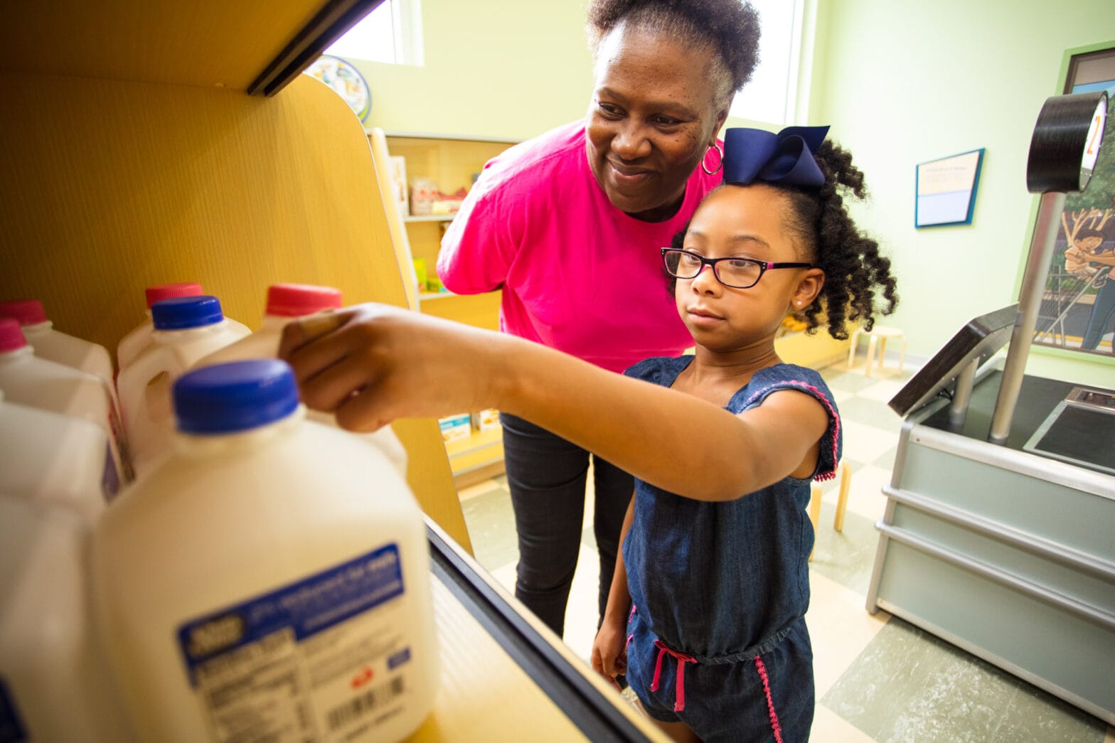 A little girl and woman reach for toy milk in the Pelican Pantry