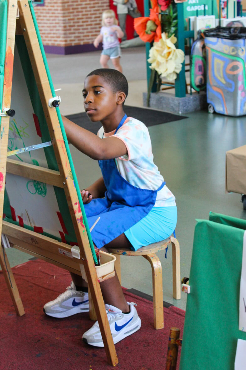 A young boy focusing on painting while sitting down in front of an art easel.