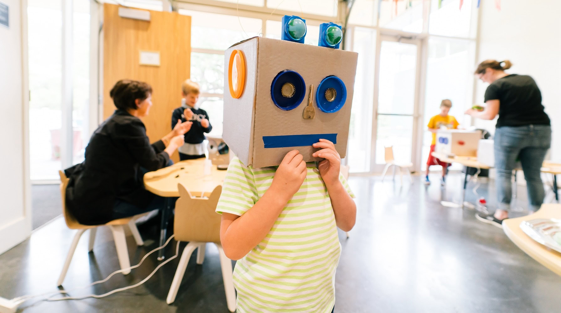 A young child with a cardboard box decorated as a robot placed on their head