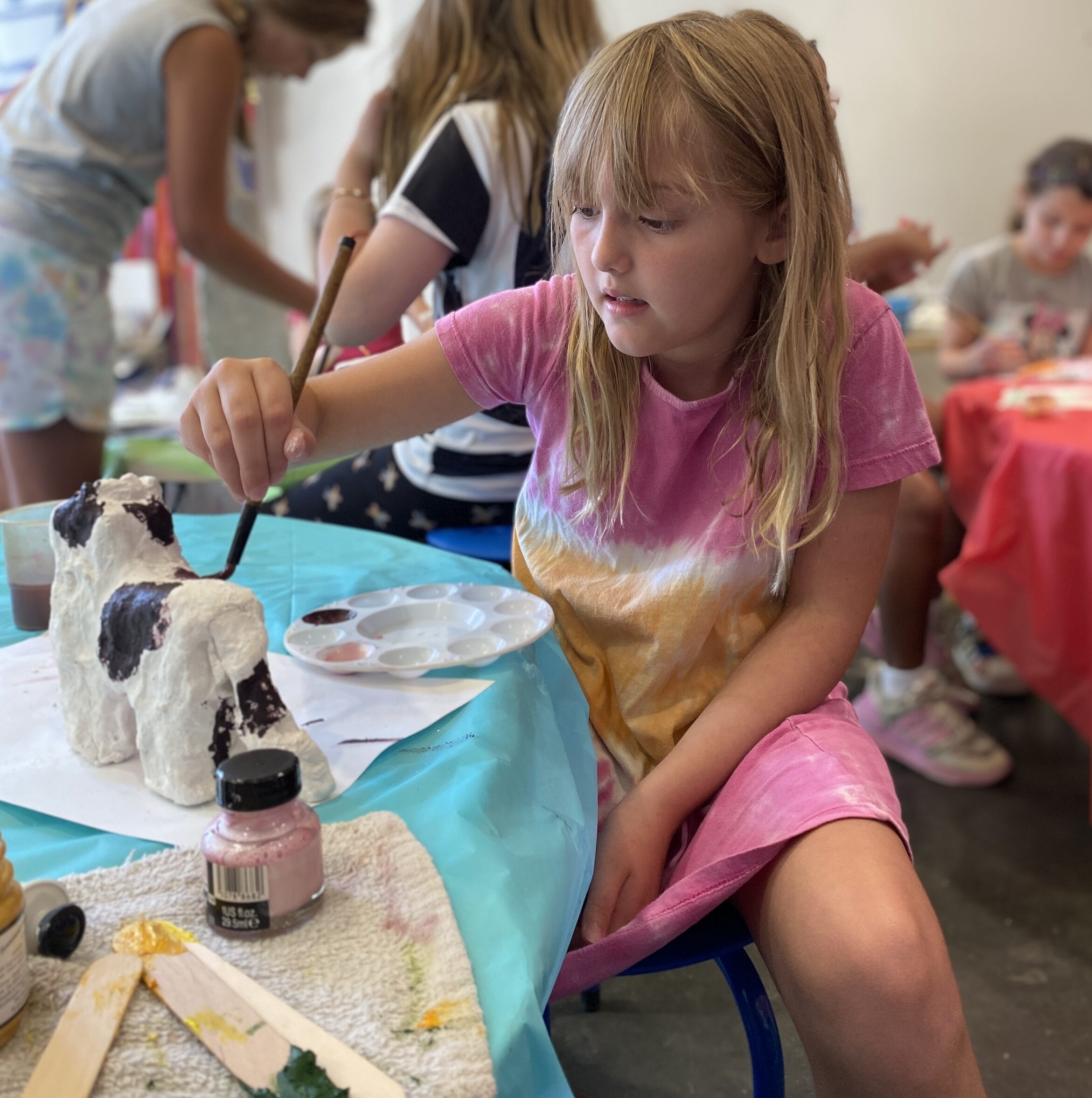 Girl wearing a pink dress sitting at a table painting a paper mache cow.